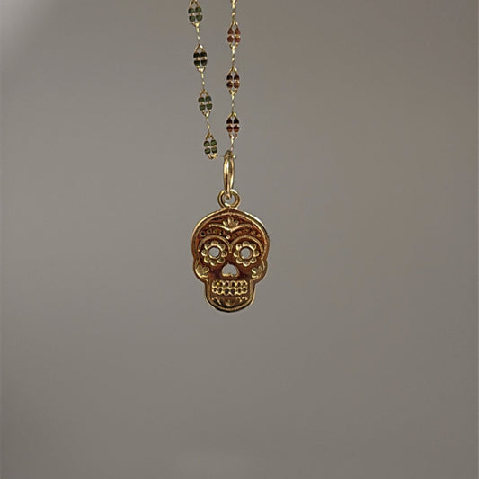 A 14k gold sugar skull charm with intricate details, suspended from a gold chain against a grey background.
