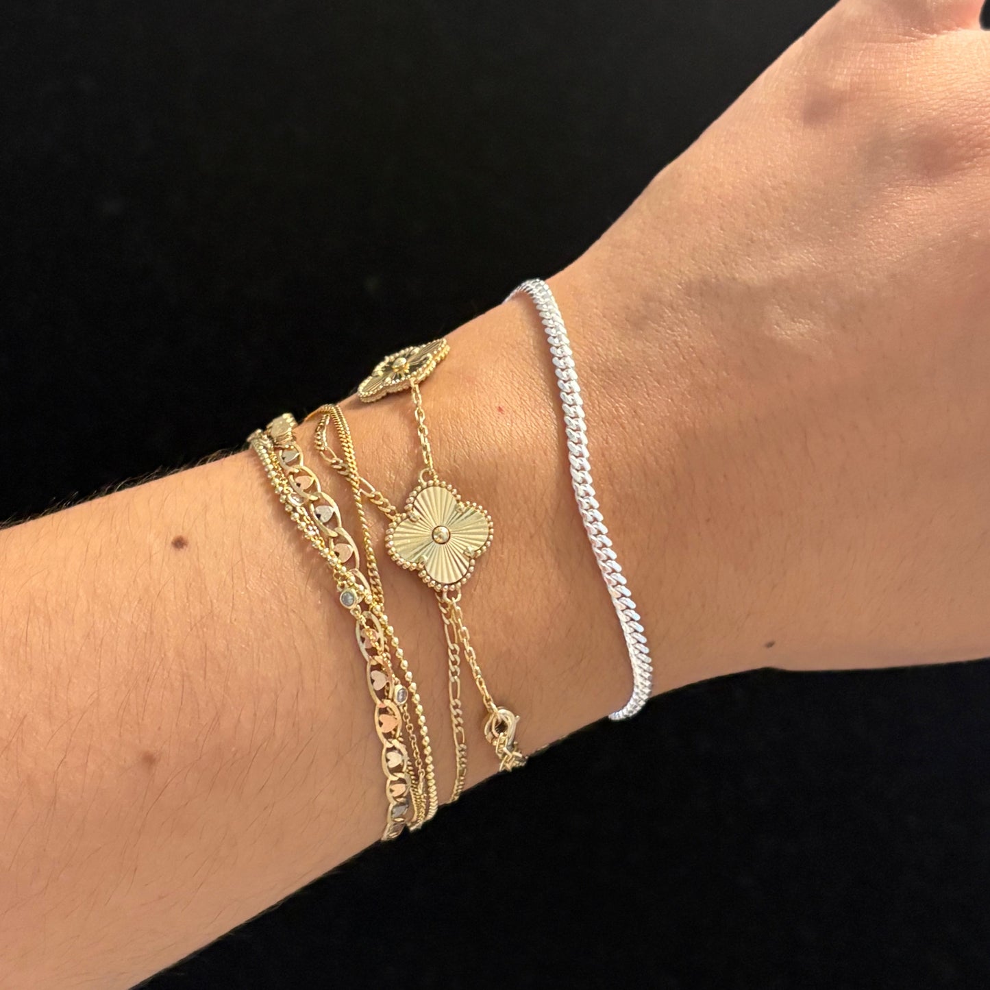 A close-up of a person's wrist wearing multiple delicate bracelets, including a silver chain bracelet, a gold chain bracelet with a flower charm, and textured gold bracelets, set against a solid black background.
