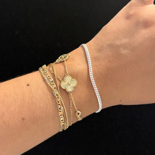 A close-up of a person's wrist wearing multiple delicate bracelets, including a silver chain bracelet, a gold chain bracelet with a flower charm, and textured gold bracelets, set against a solid black background.