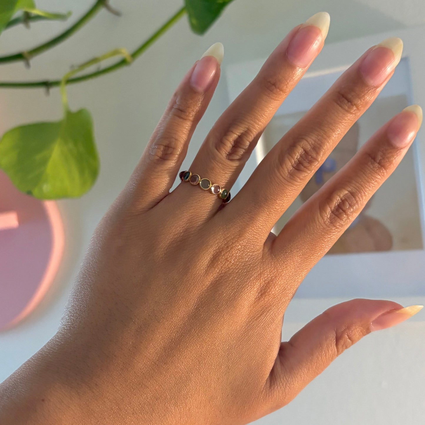 A close-up of a hand wearing a delicate gold ring with small geometric links, set against a bright, softly blurred background with green leaves.