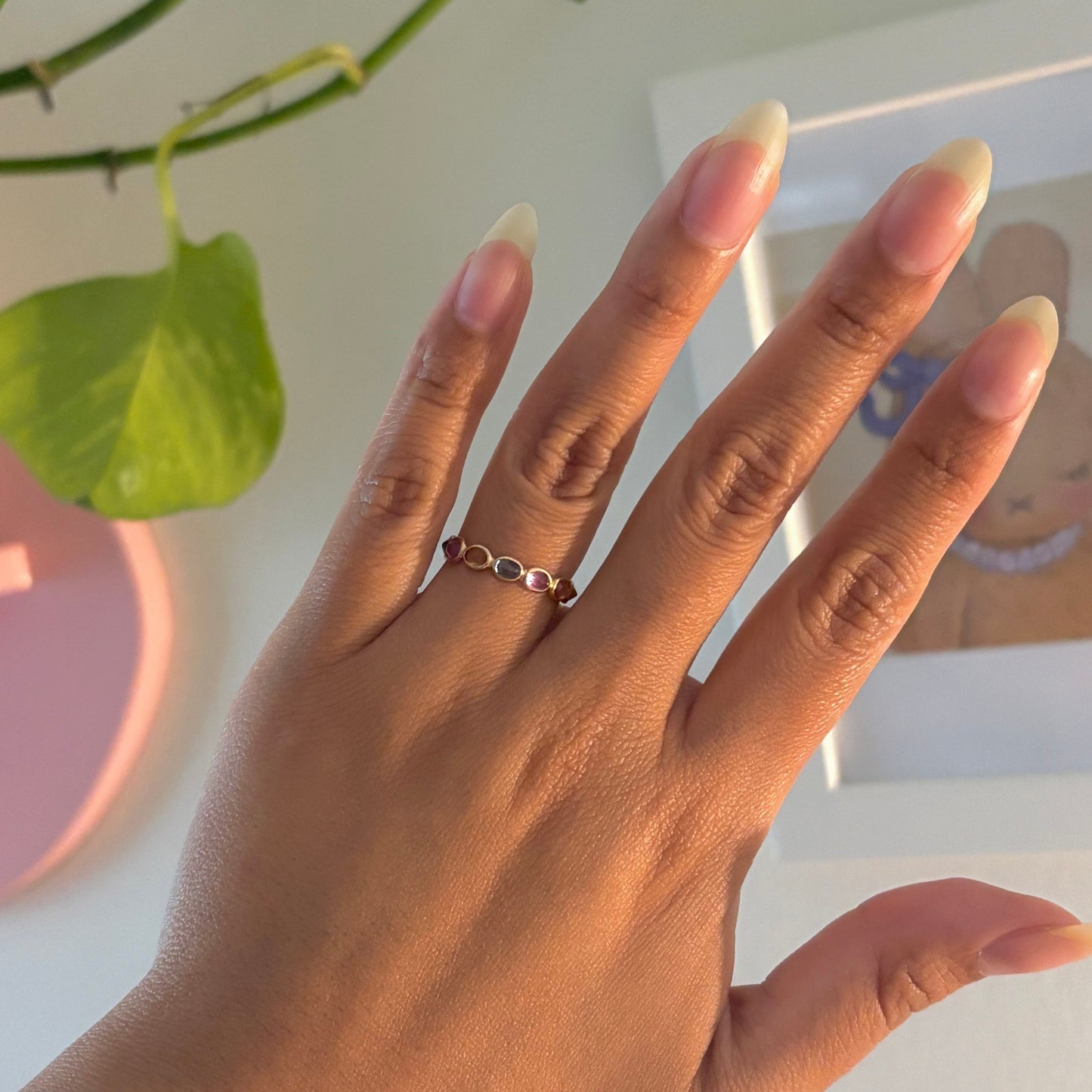 A close-up of a hand wearing a delicate silver ring with small red and white stones, set against a bright indoor background with a green plant and a white frame.