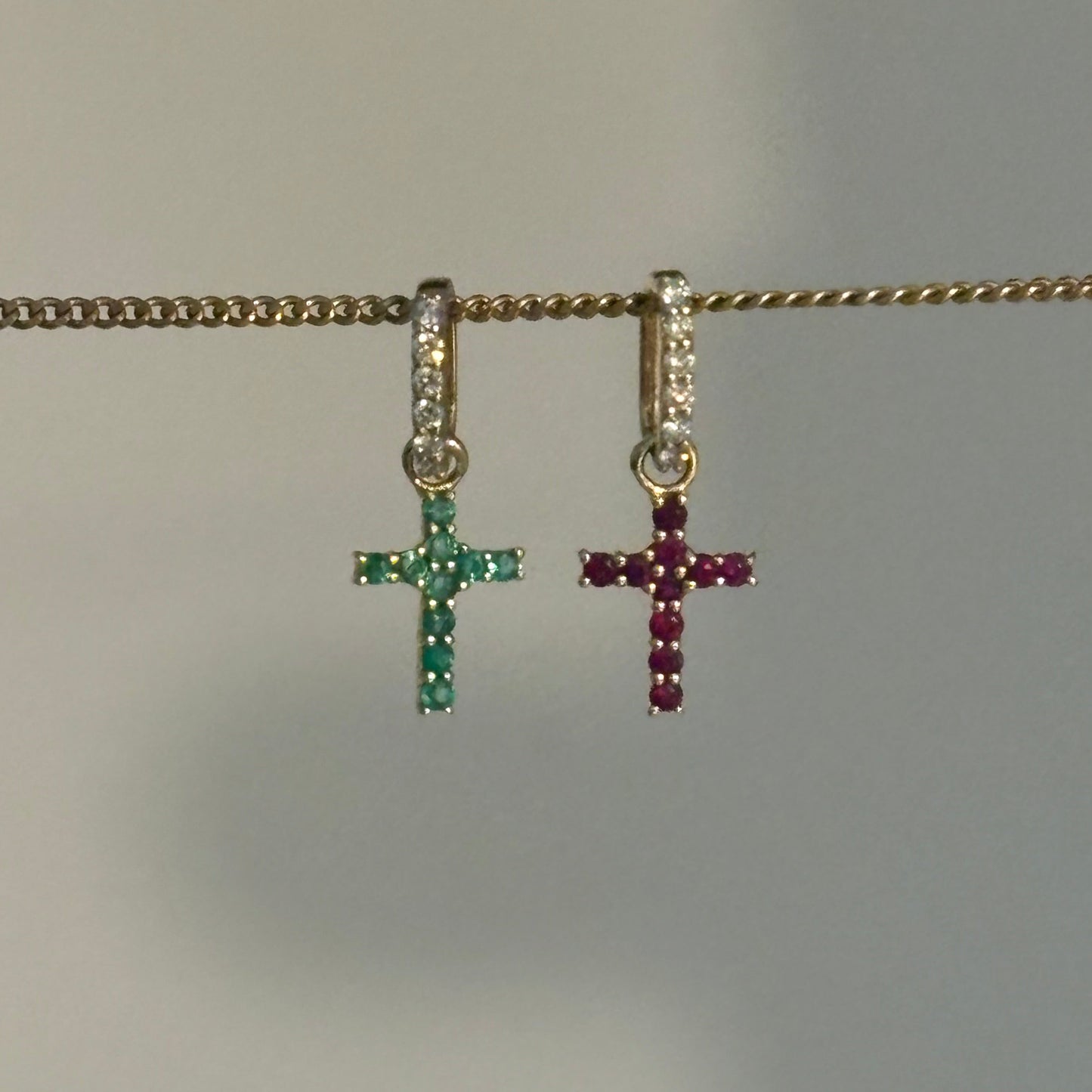 A close-up of a gold chain necklace with two small cross charms hanging vertically. The top charm is gold with dark red stones, and the bottom charm is gold with green stones.