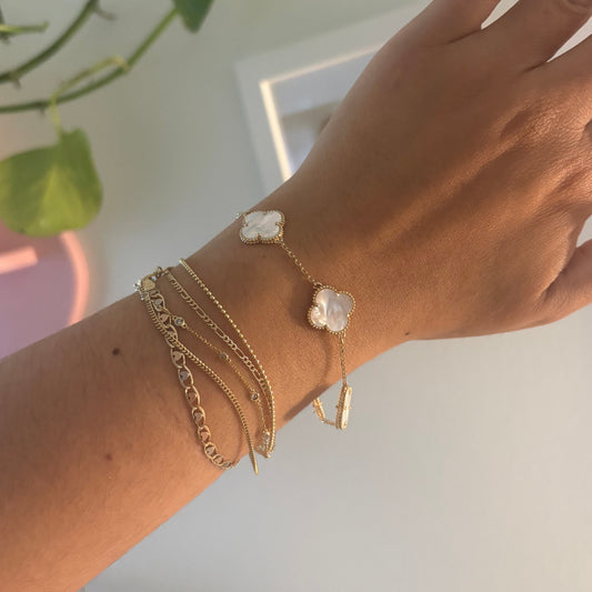 A close-up of a person's wrist wearing multiple delicate gold chain bracelets, including one with a small white flower charm and another with a gold bar, against a softly blurred indoor background with a green plant.