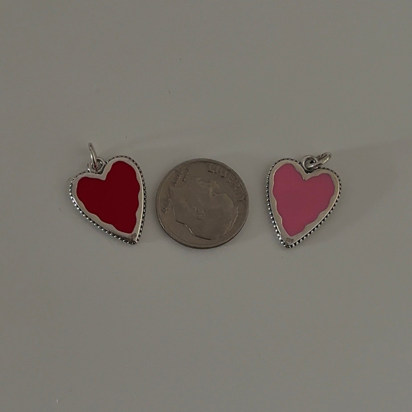 Two heart-shaped pendants, one red and one pink, with a coin for scale on a gray background.