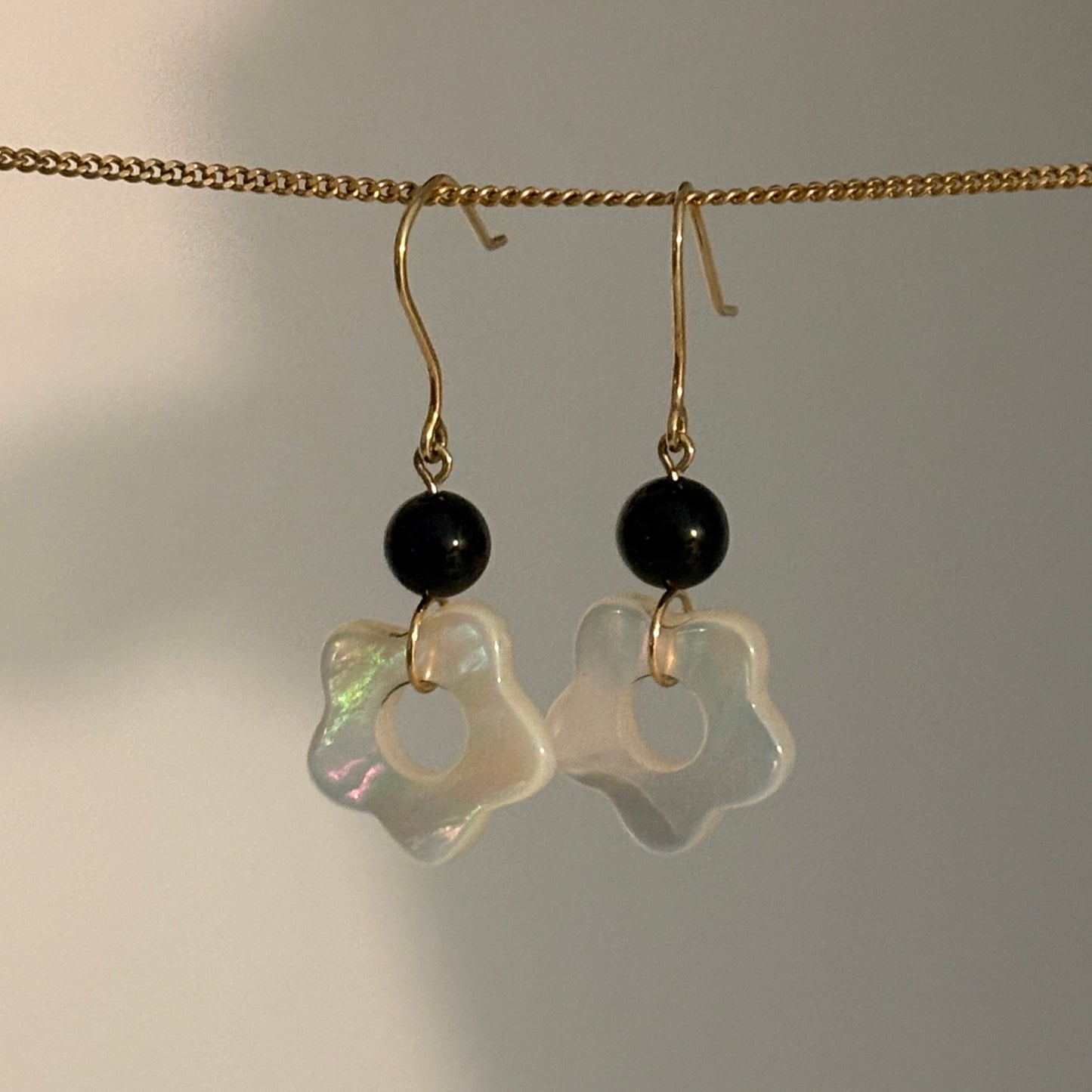A pair of dangling earrings hanging from a gold chain against a plain light background. Each earring has a small black round bead and a translucent iridescent white flower-shaped charm.