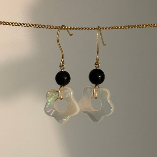 A pair of dangling earrings hanging from a gold chain against a plain light background. Each earring has a small black round bead and a translucent iridescent white flower-shaped charm.