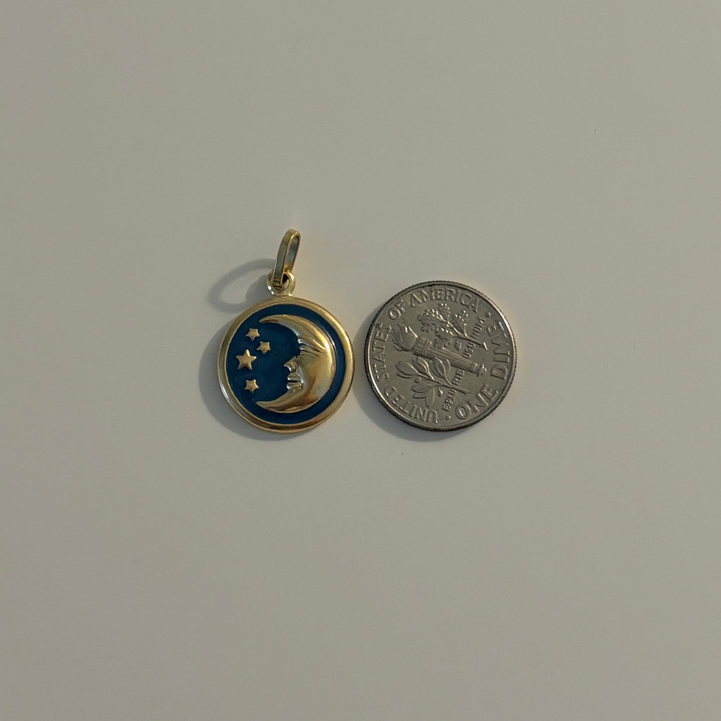 A small round pendant charm with a gold-tone bail, featuring a blue and gold crescent moon and star design, shown next to a silver coin for scale on a plain light gray background.