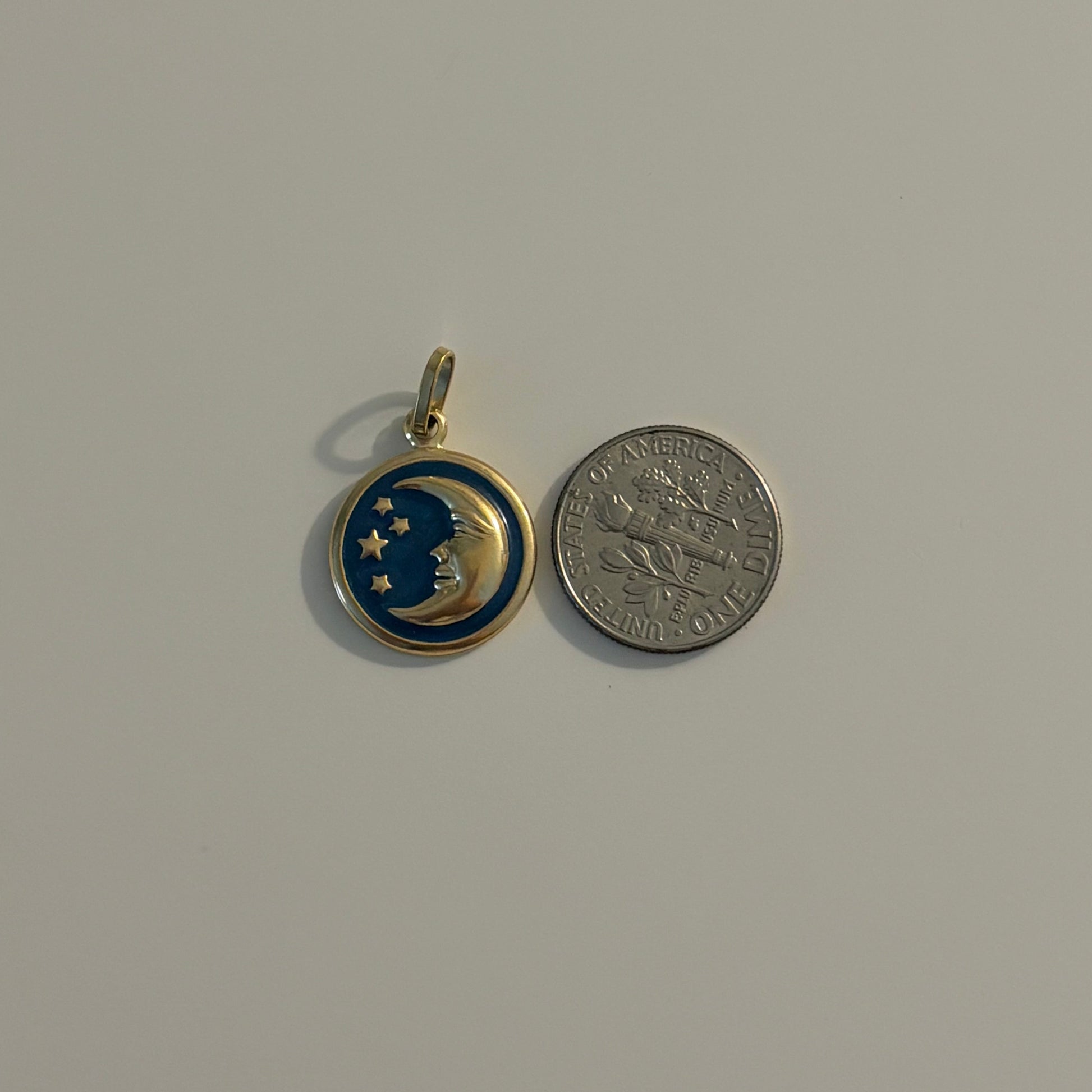 A small round pendant charm with a gold-tone bail, featuring a blue and gold crescent moon and star design, shown next to a silver coin for scale on a plain light gray background.