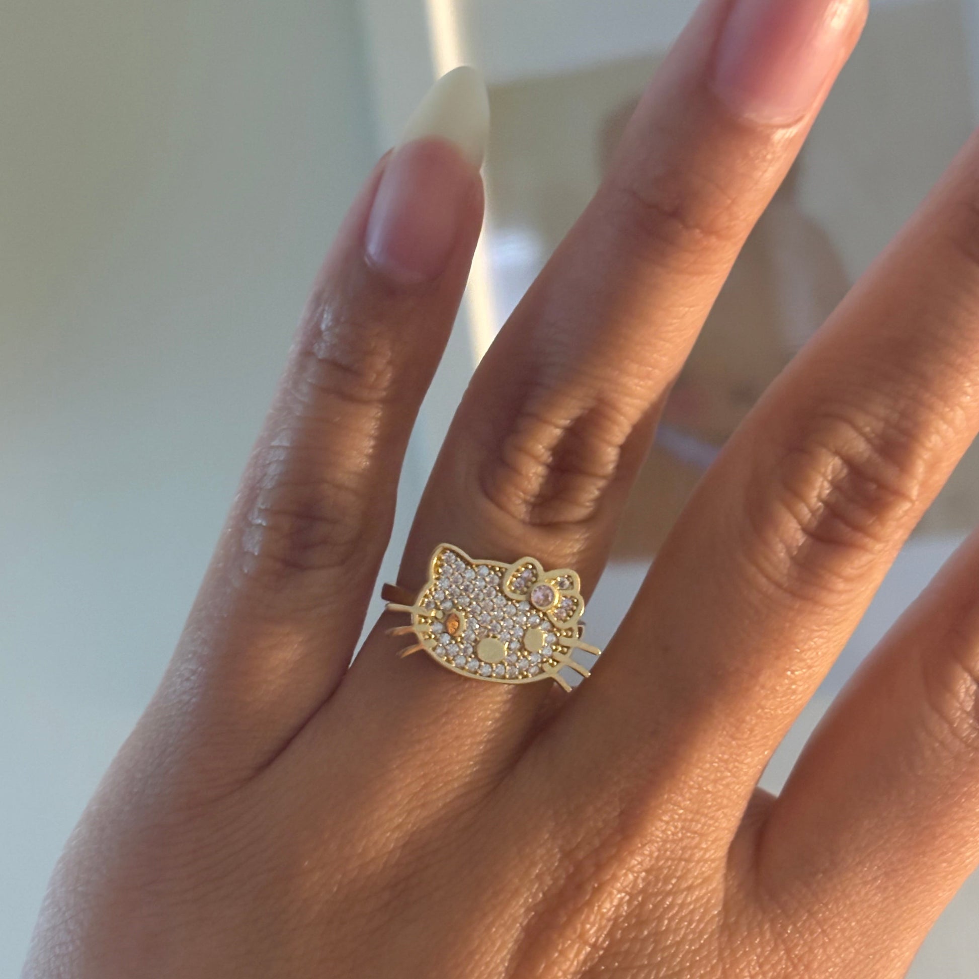 A gold ring featuring a cat face design paved with small clear stones, shown worn on a person's finger.