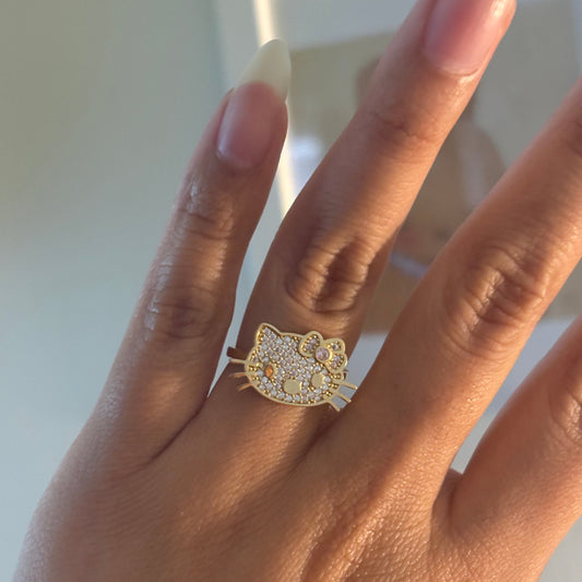A gold ring featuring a cat face design paved with small clear stones, shown worn on a person's finger.