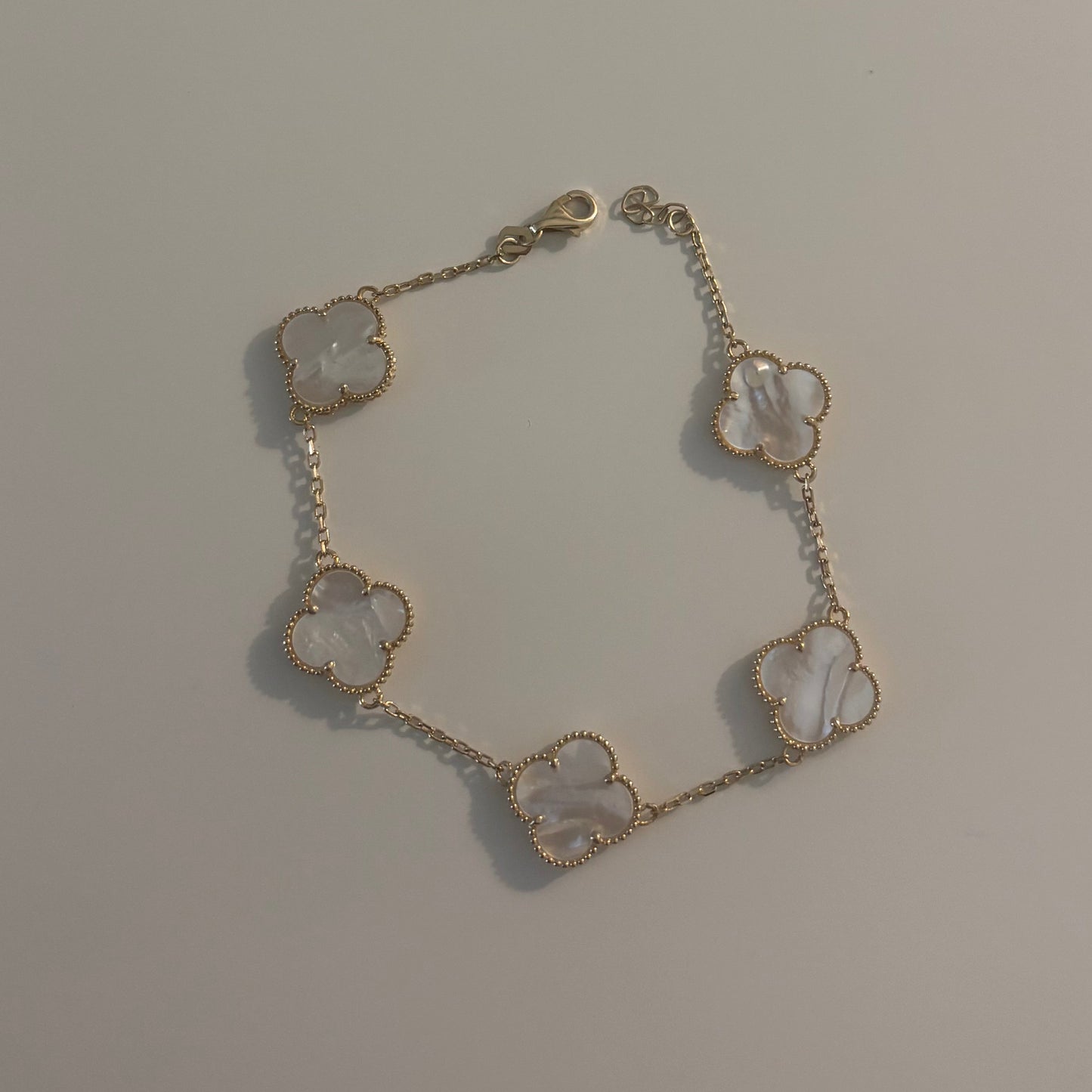 A delicate bracelet featuring five small clover-shaped charms with white mother-of-pearl inlays, set in a gold-tone chain, displayed on a plain light gray surface.