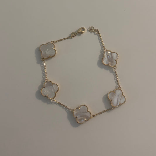 A delicate bracelet featuring five small clover-shaped charms with white mother-of-pearl inlays, set in a gold-tone chain, displayed on a plain light gray surface.