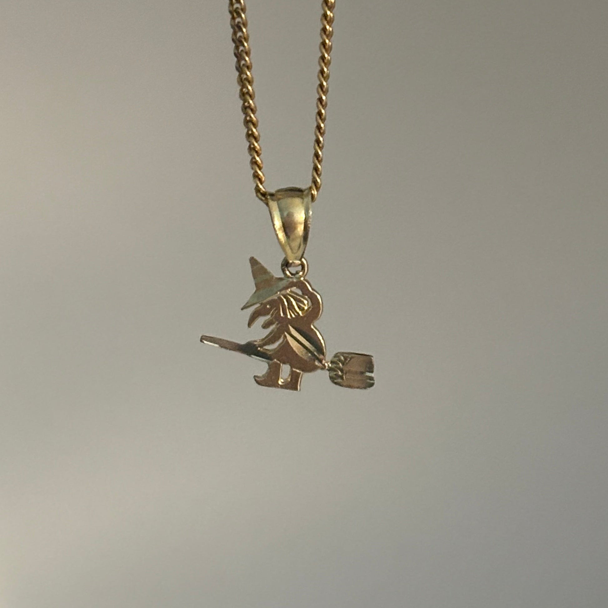 A gold chain necklace with a small witch riding a broomstick pendant, shown against a plain light gray background.