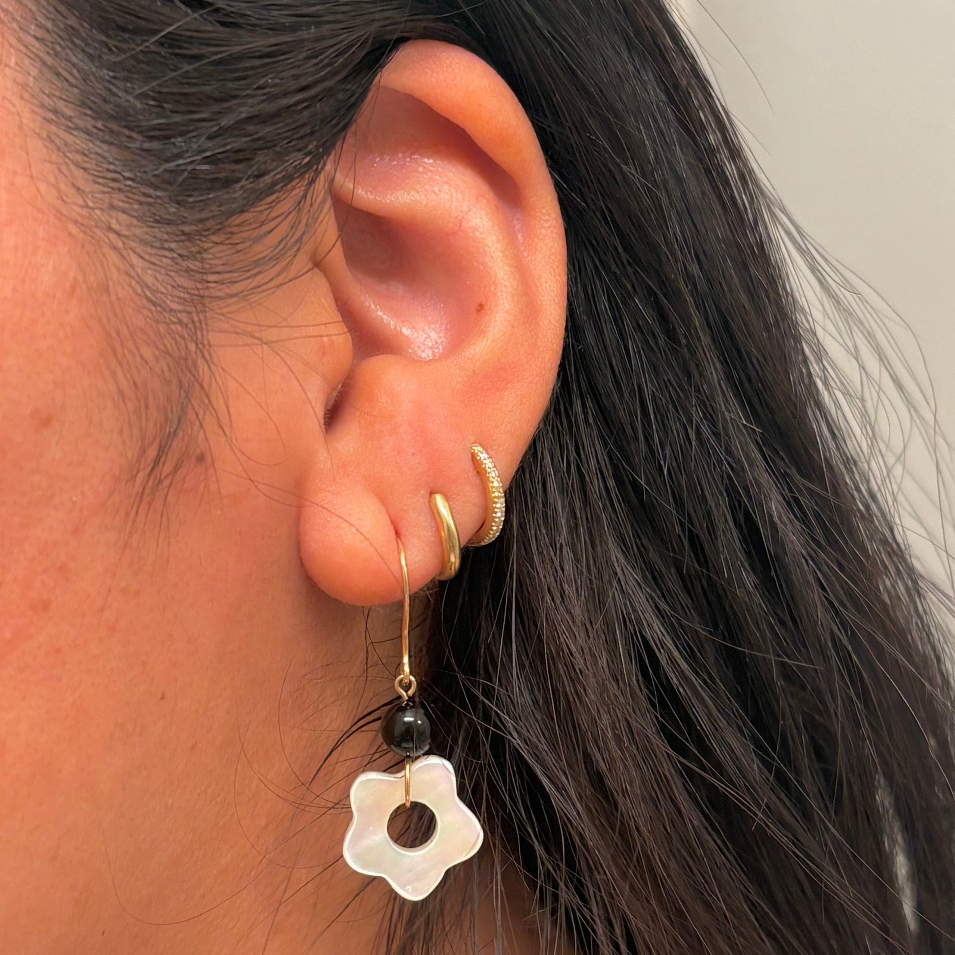 A close-up side view of a person's ear wearing a gold hoop earring with a dangling white flower charm and a small black bead.
