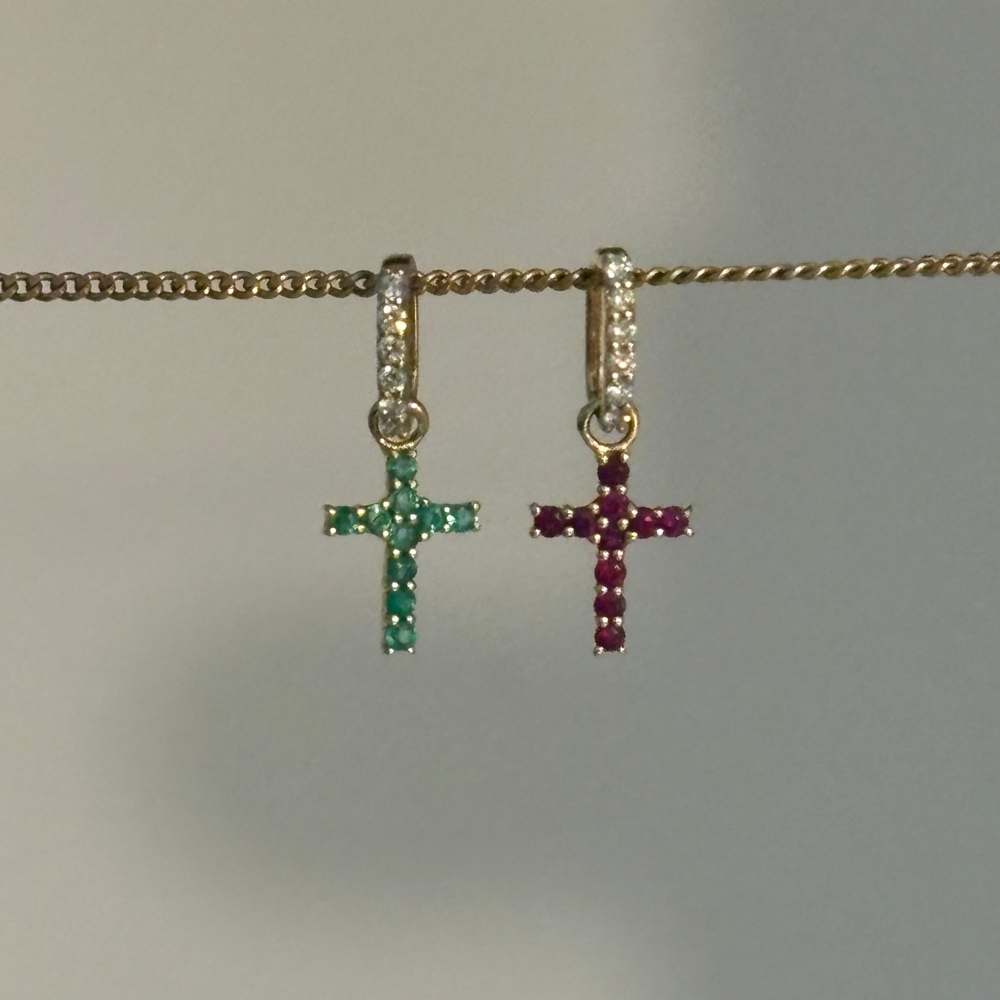 A close-up of a gold chain necklace with two small cross charms hanging vertically. The top charm is gold with dark red stones, and the bottom charm is gold with green stones.