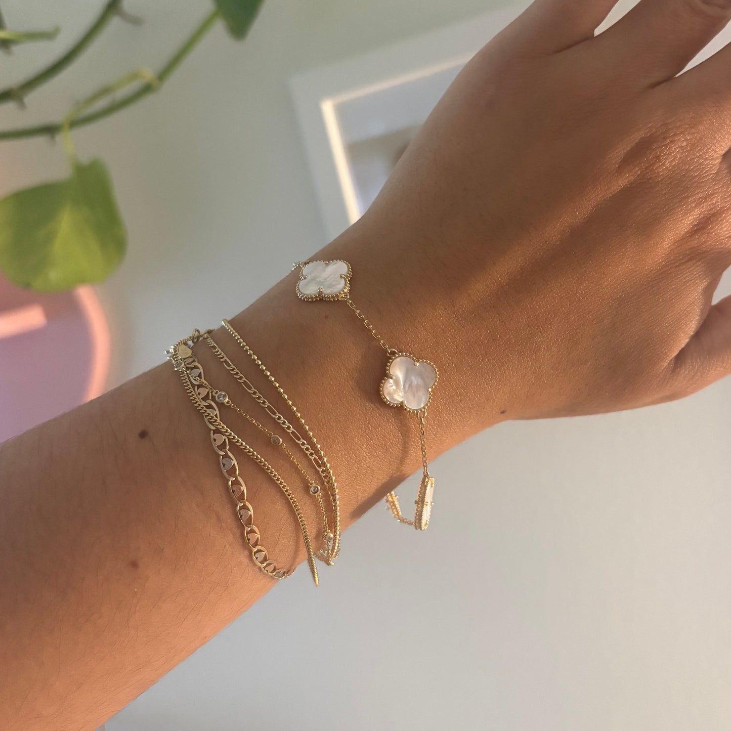 A close-up of a person's wrist wearing multiple delicate gold chain bracelets, including one with a small white flower charm and another with a gold bar, against a softly blurred indoor background with a green plant.