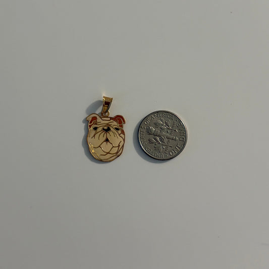 A small gold pendant shaped like a bulldog's face with red and black enamel details, shown next to a silver coin for scale on a plain light gray background.