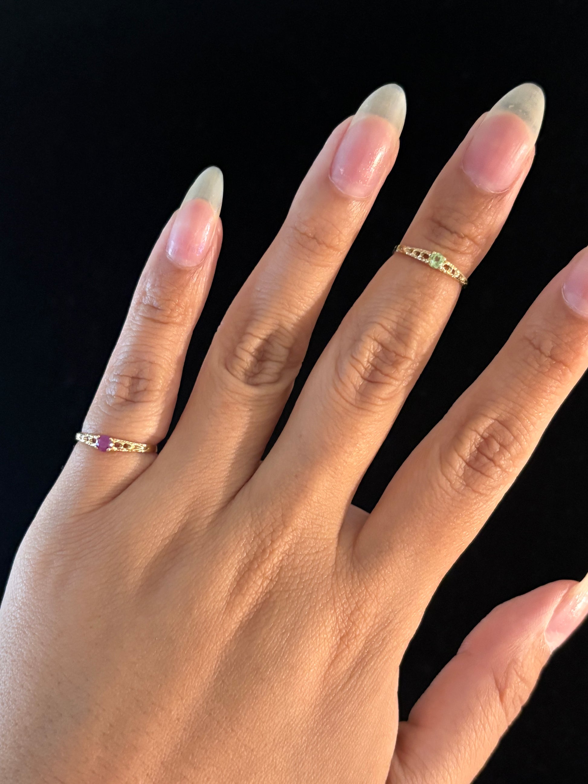 A close-up of a hand wearing two delicate gold rings with small colored gemstones, set against a solid black background.
