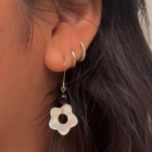 A close-up of a person's ear wearing multiple earrings, including a white flower-shaped drop earring with a black bead and gold hook, along with small gold hoop earrings.