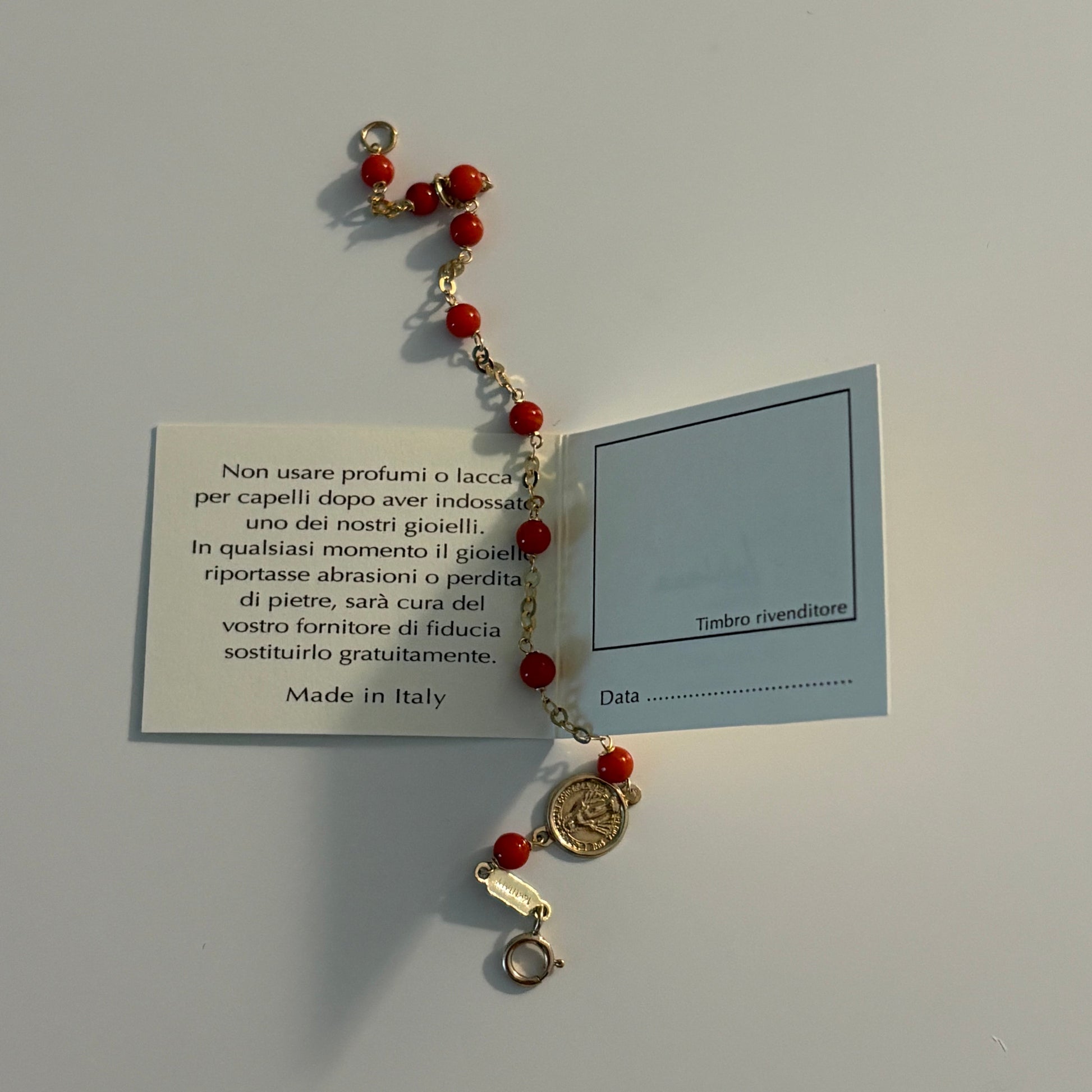 A small beaded bracelet with red beads, gold-toned metal links, and a round gold coin charm, displayed on a light gray background with a small printed card behind it.