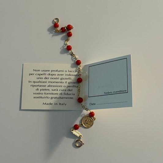 A small beaded bracelet with red beads, gold-toned metal links, and a round gold coin charm, displayed on a light gray background with a small printed card behind it.