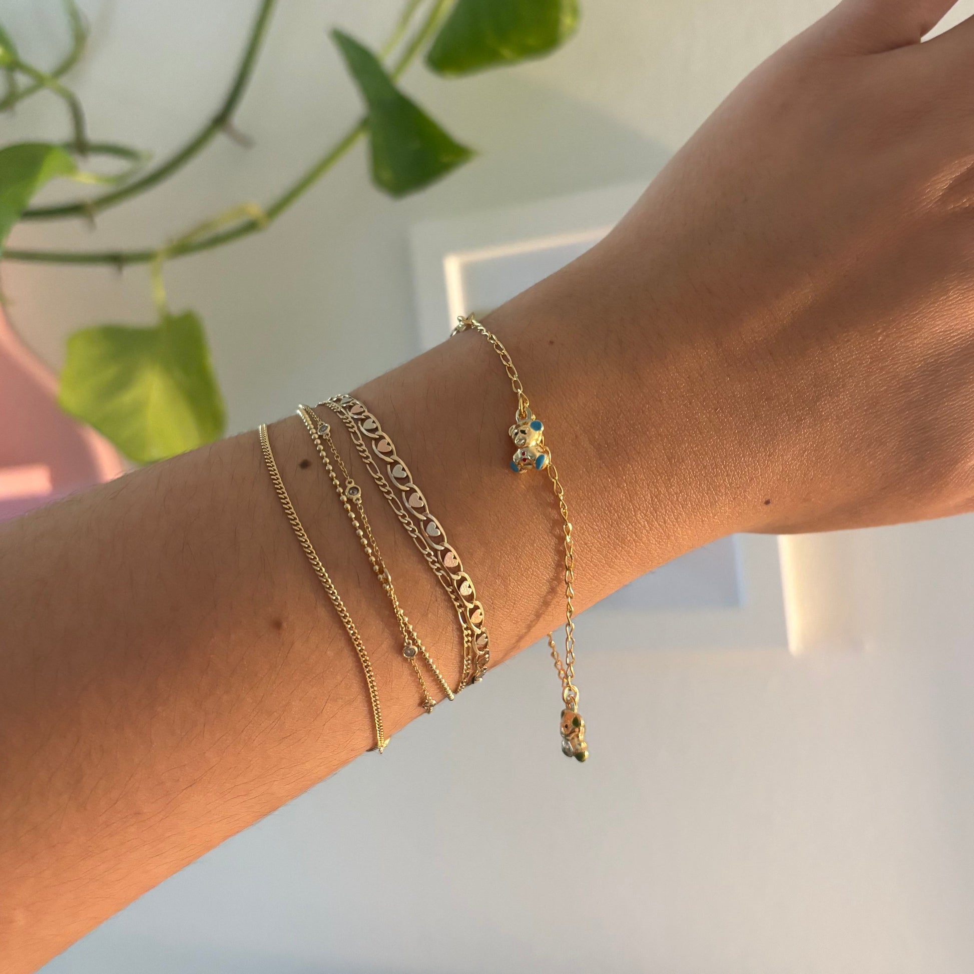 A close-up of a person's wrist wearing multiple delicate gold bracelets, including a thin chain bracelet with a small clear stone and stacked textured chain bracelets, set against a softly blurred indoor background with a green plant.