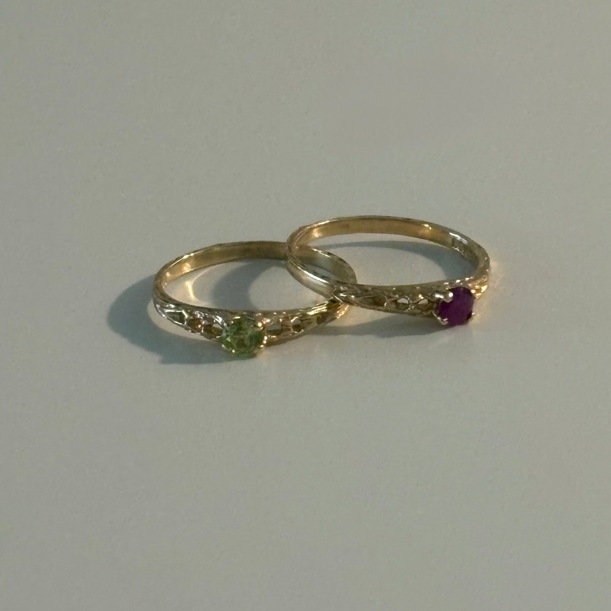 Two gold rings with small gemstones, one featuring a green stone and the other a purple stone, displayed on a light gray surface.
