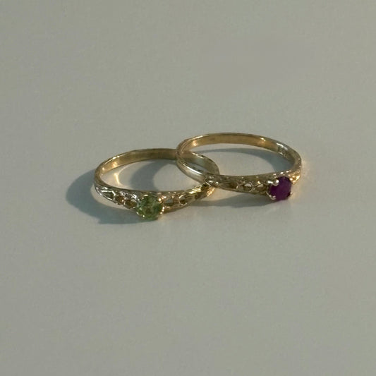 Two gold rings with small gemstones, one featuring a green stone and the other a purple stone, displayed on a light gray surface.