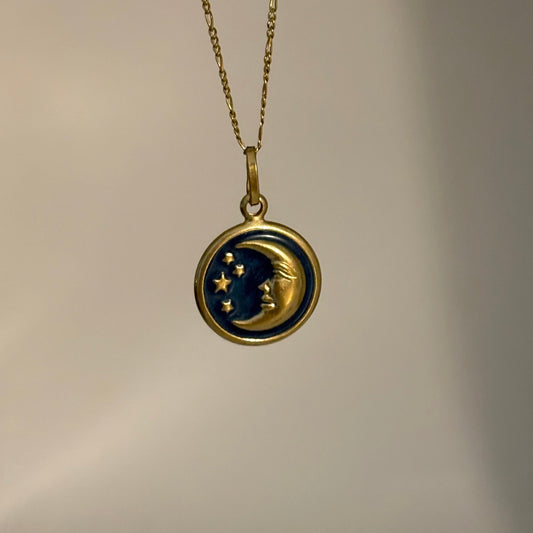 A gold-toned pendant necklace featuring a crescent moon and stars design in dark blue and gold, hanging against a plain light gray background.