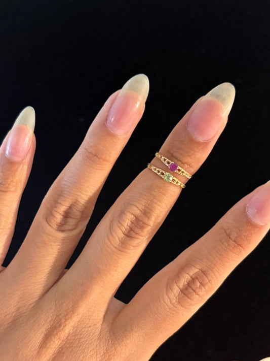 A close-up of a hand with long, almond-shaped nude nails wearing two delicate gold rings. One ring features a small round pink gemstone, and the other features a small round green gemstone, both accented with tiny clear stones, set against a solid black background.