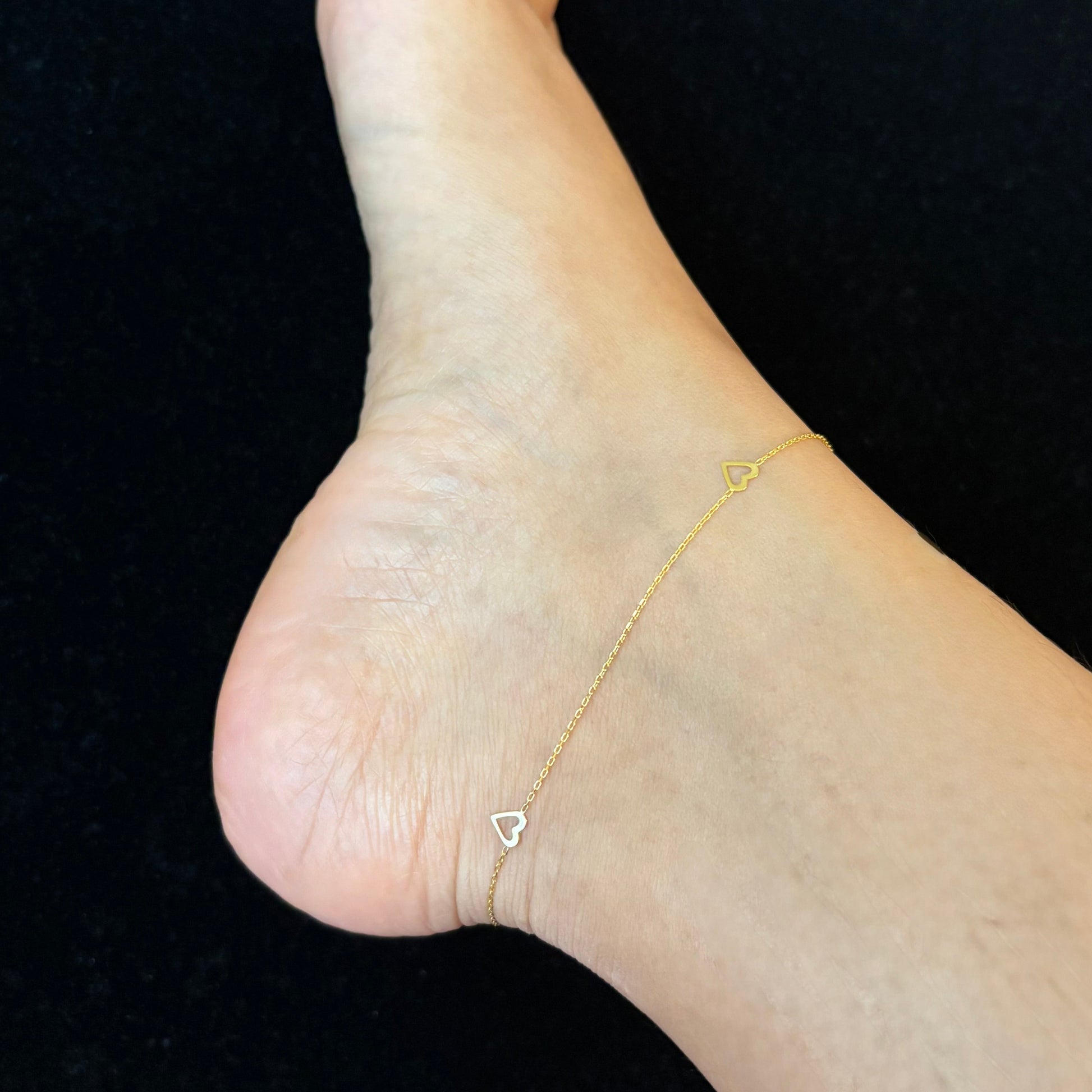 Foot wearing a gold anklet with a small heart charm on a black surface.