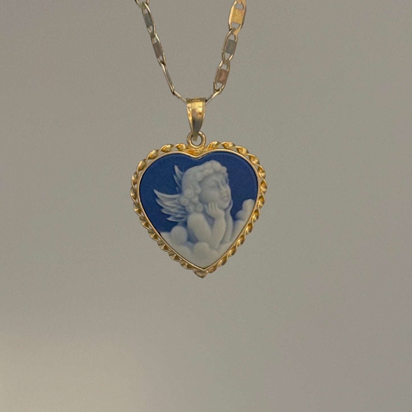 Heart-shaped pendant with angel design on a gold chain against a gray background