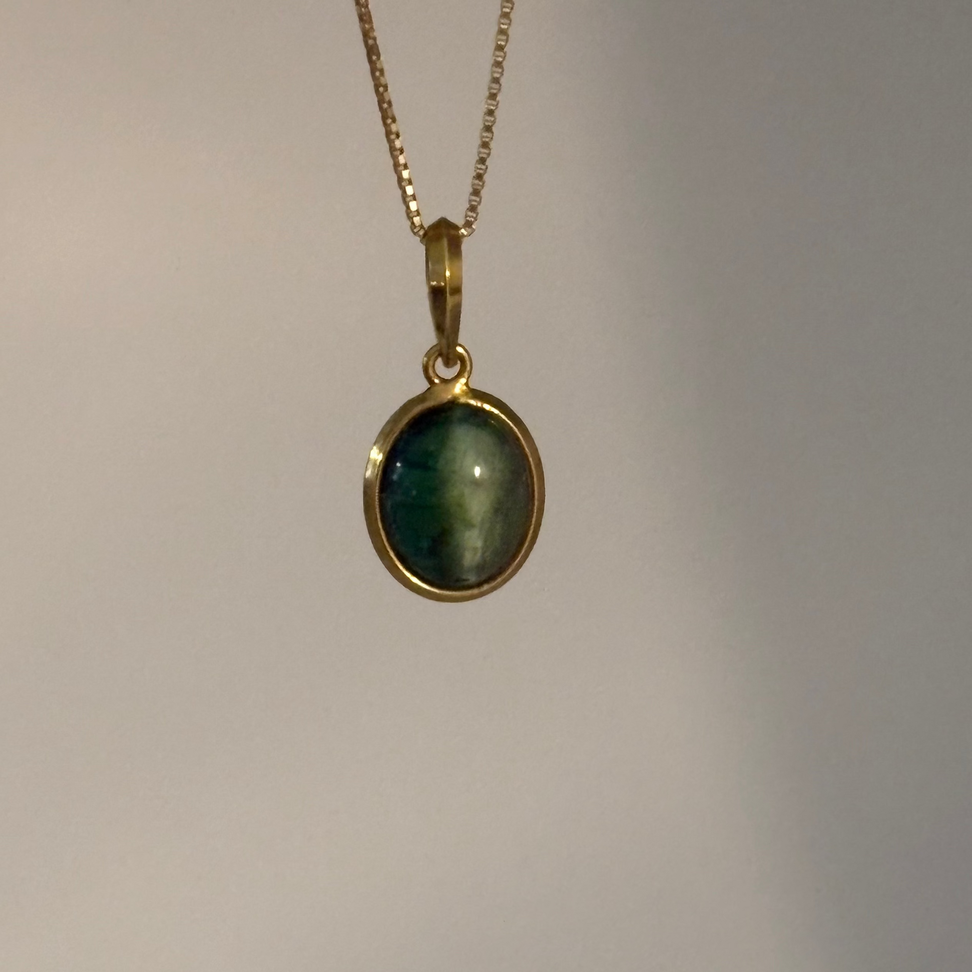 A gold chain necklace with an oval pendant featuring a dark green stone with a subtle sheen, set in a gold bezel, shown against a plain light gray background.