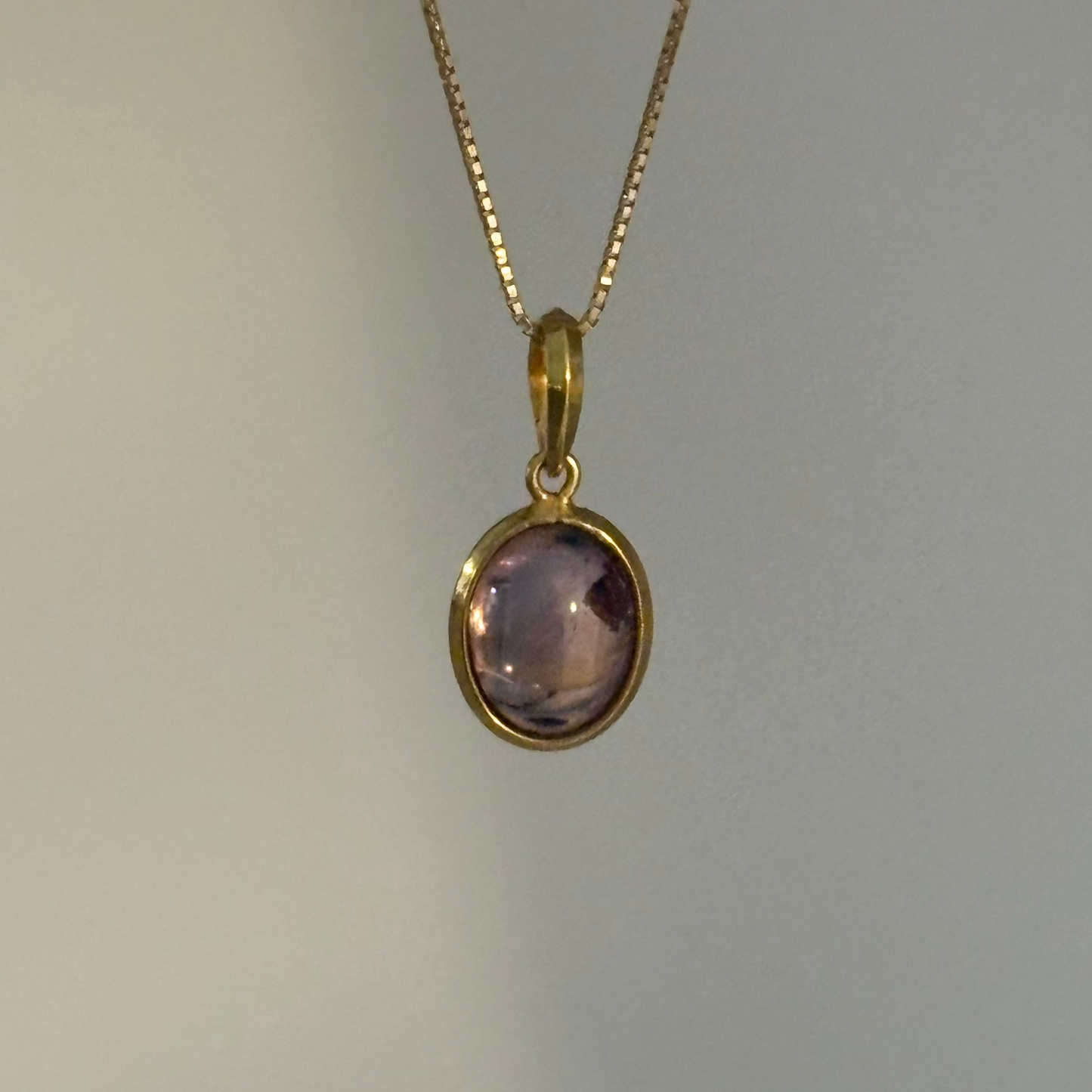 A gold chain necklace with an oval bezel-set light purple amethyst pendant, shown against a plain light gray background.
