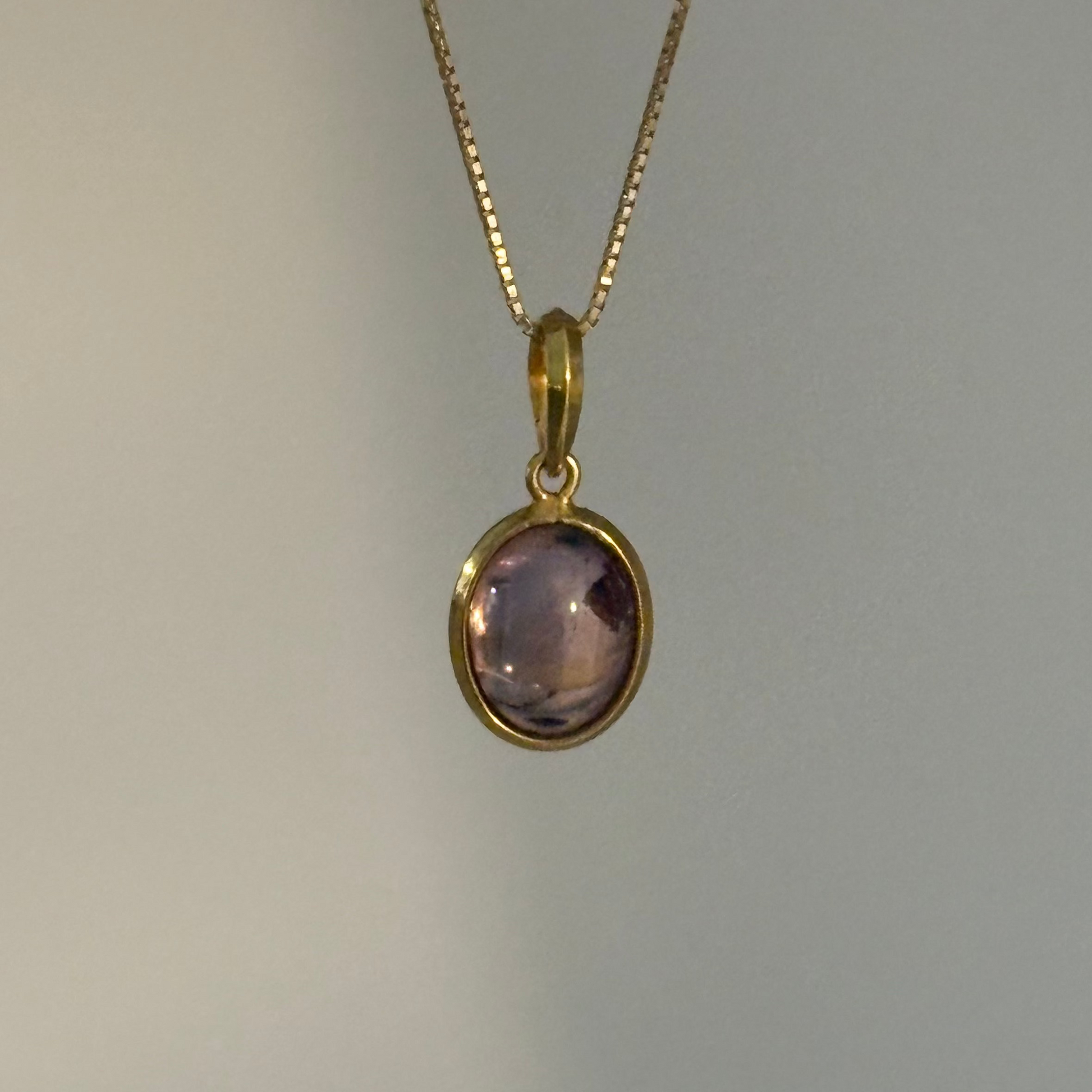 A gold chain necklace with an oval bezel-set light purple amethyst pendant, shown against a plain light gray background.