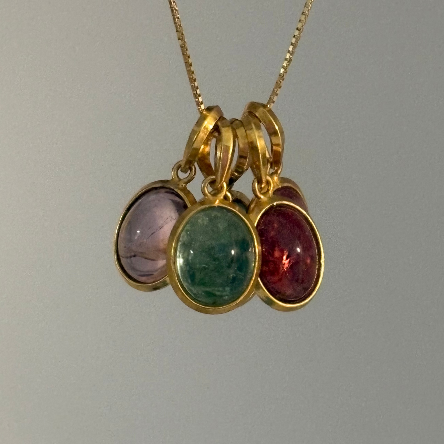 A gold chain necklace with a cluster of three oval gemstone pendants in gold bezels, featuring light purple, green, and deep red stones, shown against a plain light gray background.