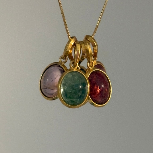 A gold chain necklace with a cluster of three oval gemstone pendants in gold bezels, featuring light purple, green, and deep red stones, shown against a plain light gray background.