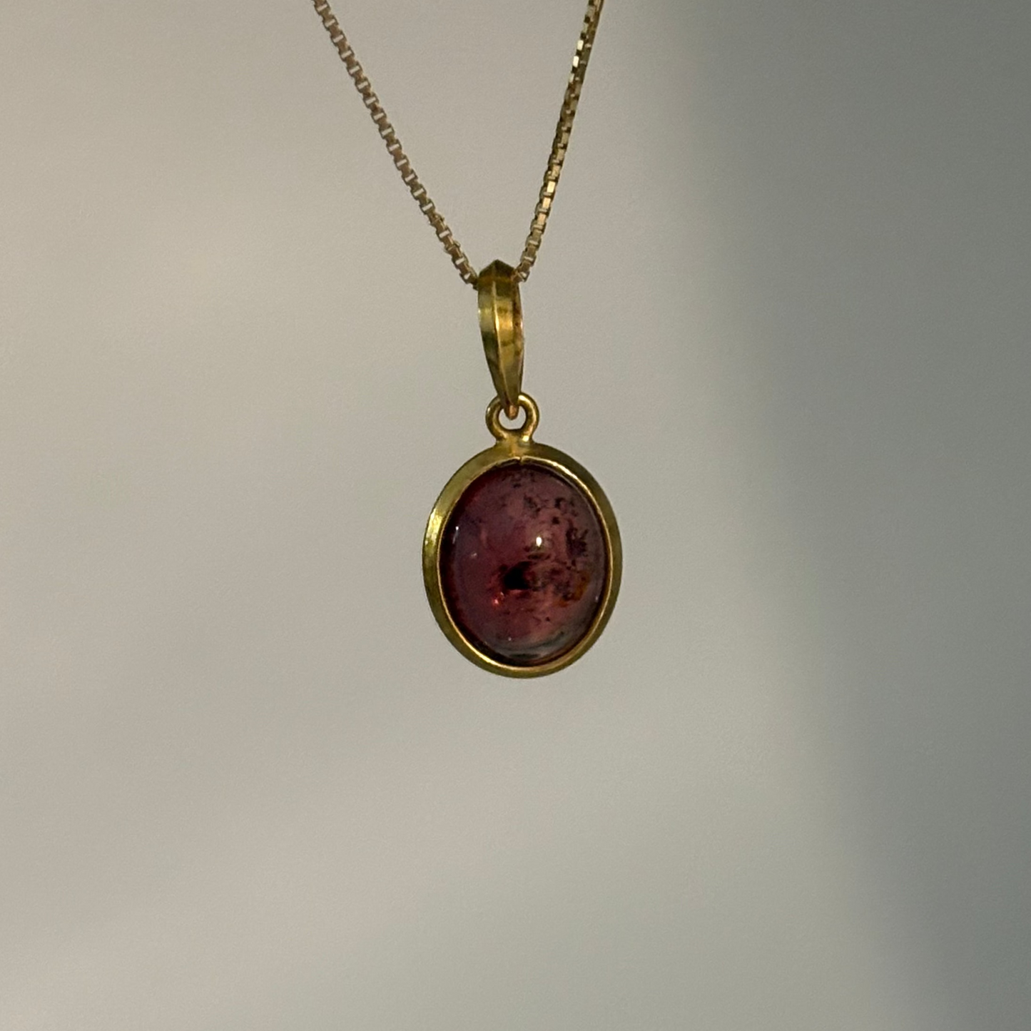 A gold chain necklace with an oval pendant featuring a pinkish-red textured stone in a gold bezel setting, shown against a neutral gray background.