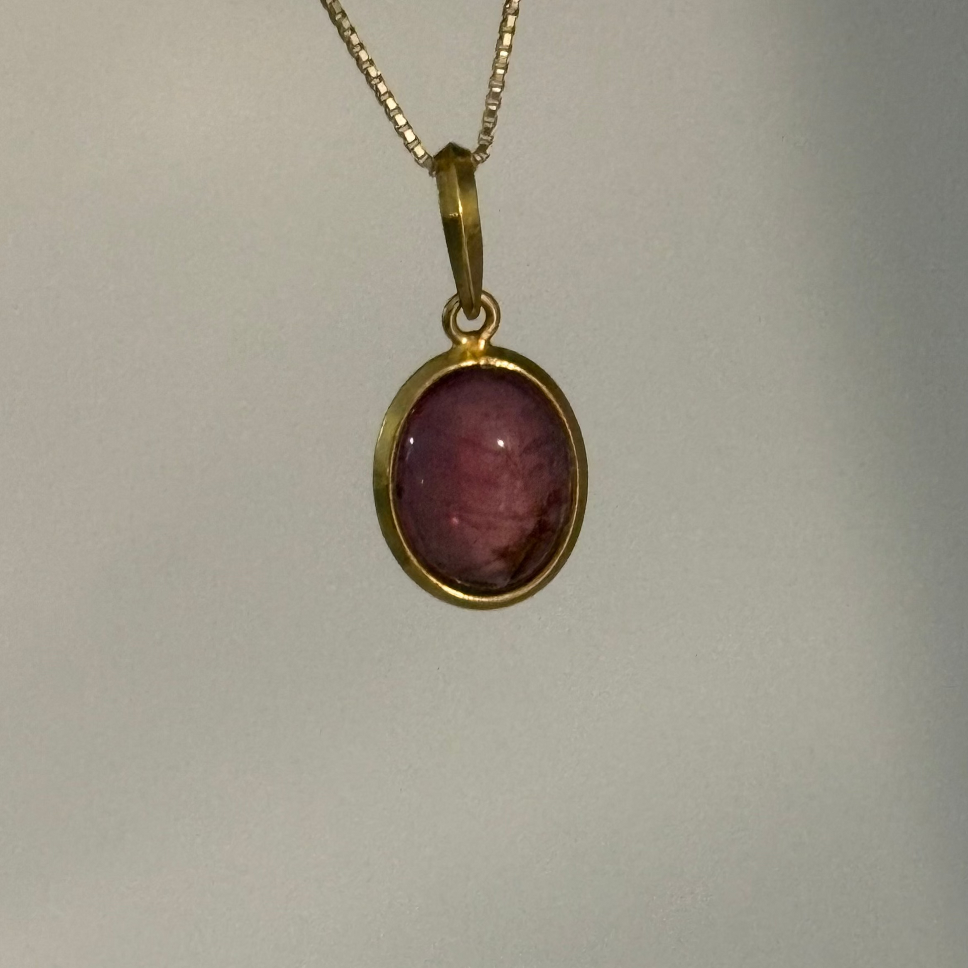 A gold chain necklace with an oval pendant featuring a pinkish-purple stone set in a gold bezel, shown against a plain light gray background.