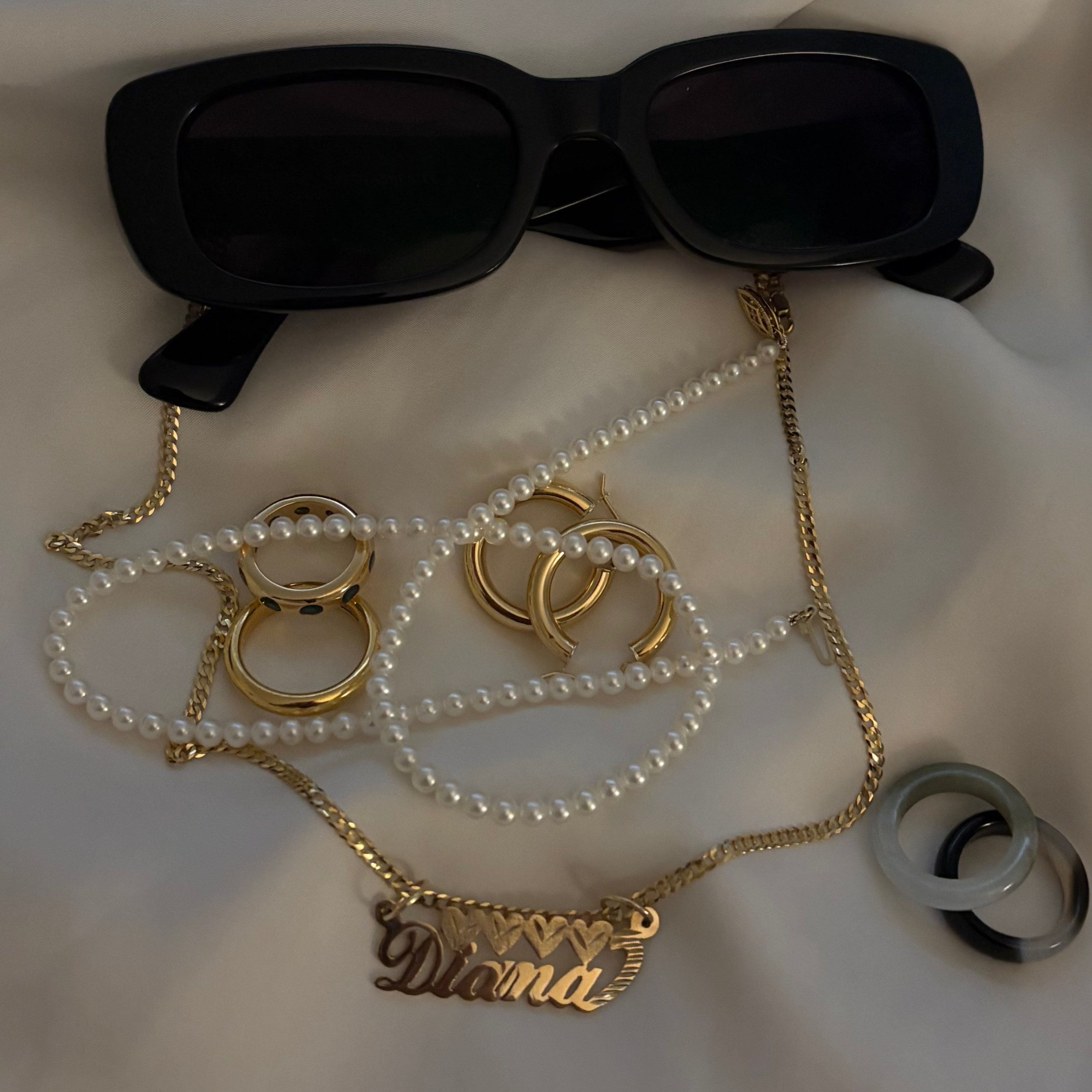 Black sunglasses and gold jewelry with 'Diana' pendant on a white fabric background